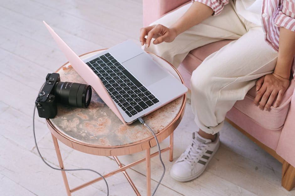 A woman dressed in casual attire sits on a pink chair working on a laptop placed on a round marble table. Next to her laptop is a professional camera connected to the device, suggesting she might be transferring media files or working on photography-related tasks.