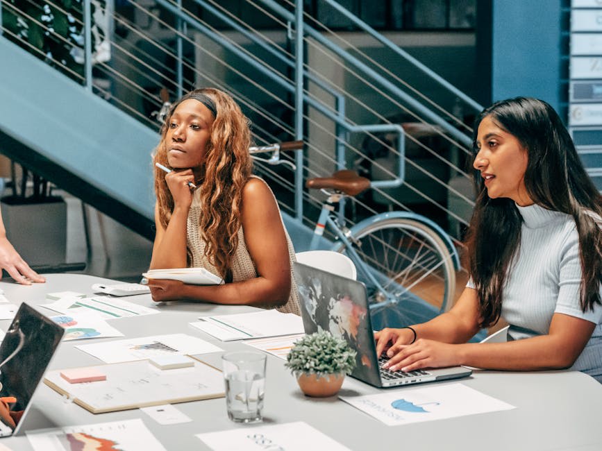 Two women sitting at a desk in an office setting, engaged in a meeting. One woman is taking notes with a pen and notepad, while the other types on a laptop. The background features a staircase and a bicycle, suggesting a modern and casual office environment. Various documents and a small plant are visible on the table, enhancing the office atmosphere.