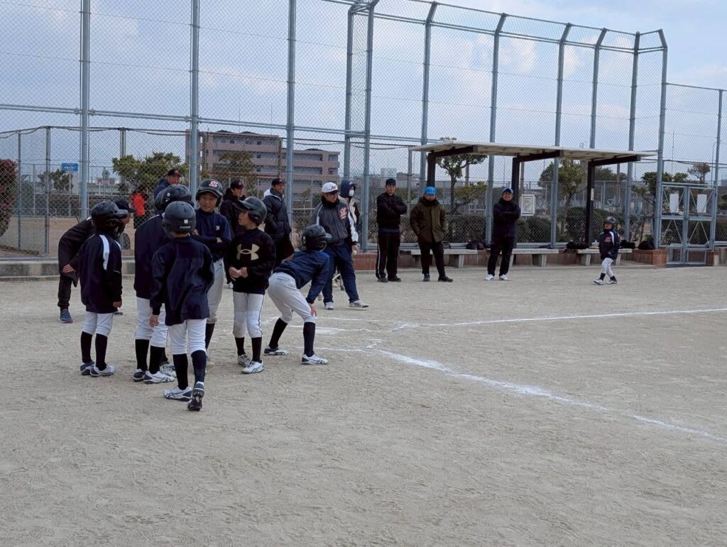 赤い服の講師が少年野球審判講習会で実技指導。ホームベース周りに集まった参加者たちが熱心に説明を聞いている。