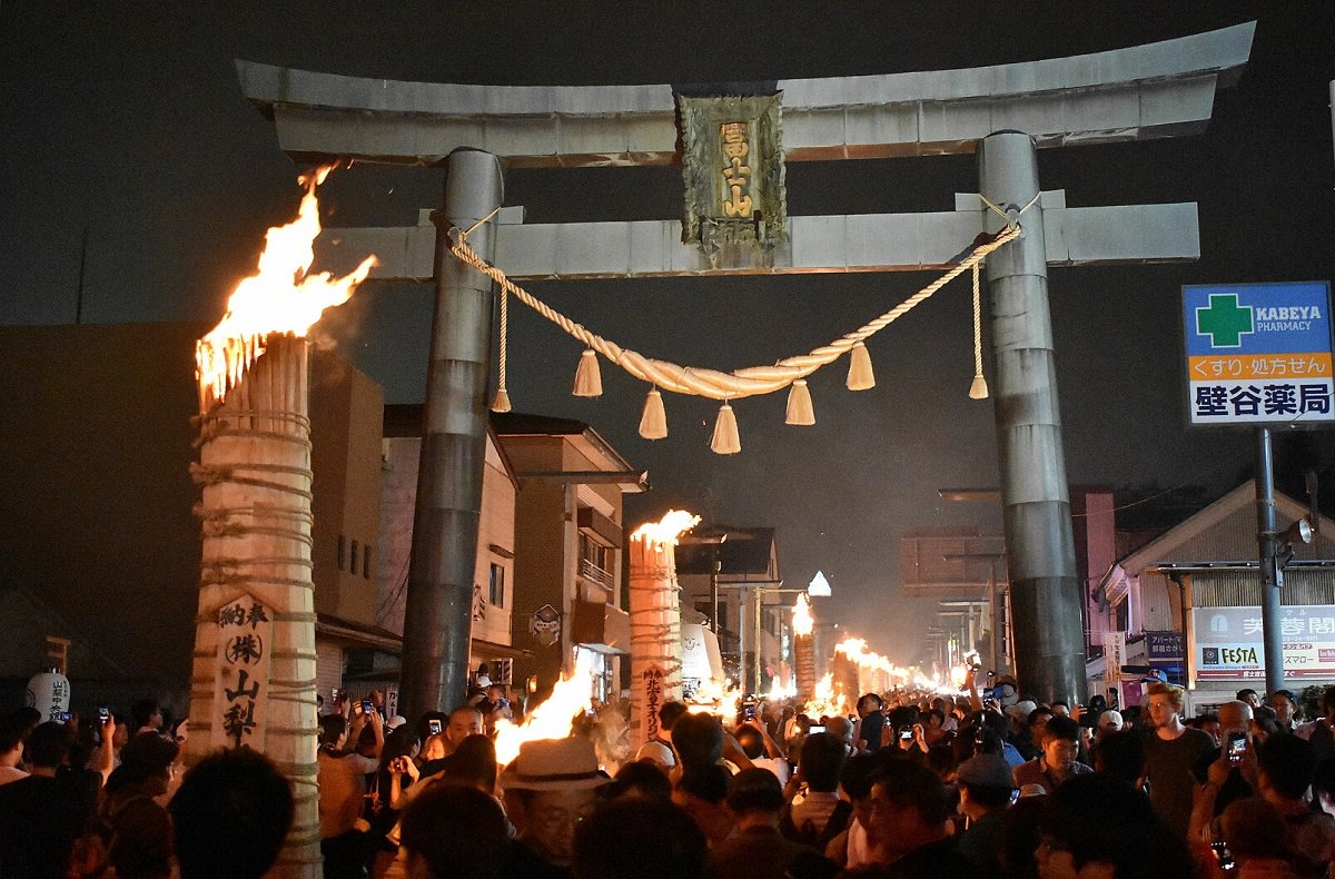 A towering, cone-shaped torch blazes brightly at the Yoshida Fire Festival, illuminating a nighttime street and crowds.