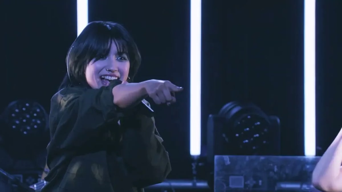 白い服を着た黒髪の女性アーティスト。ネクストブレイク候補として、紫とオレンジの光を背景にまっすぐな眼差しを向ける。