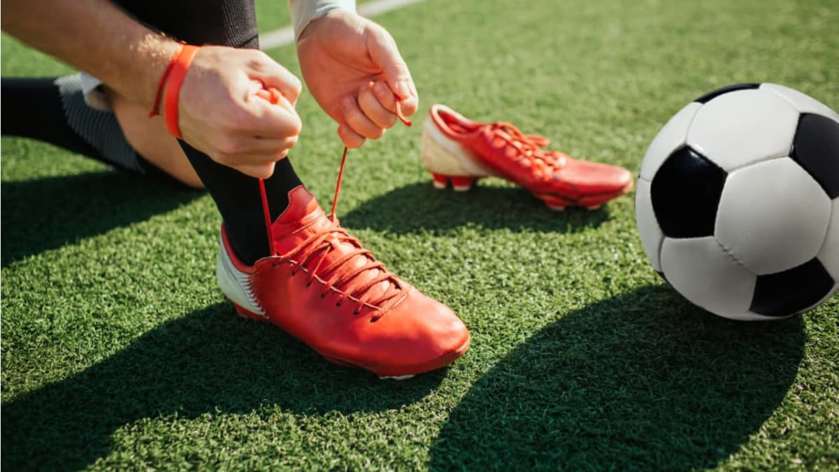 赤いユニフォームの少年が緑のサッカー場で座り、試合に向けて自分の足に合った黄色のサッカースパイクの紐をしっかりと結んでいる。