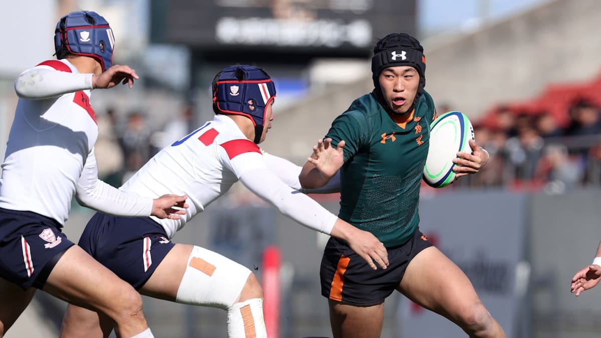 赤と白のジャージのラグビー選手がボールを抱え、敵陣へ力強く突進。全国高校ラグビーの試合日程で展開される熱いプレーを象徴する。