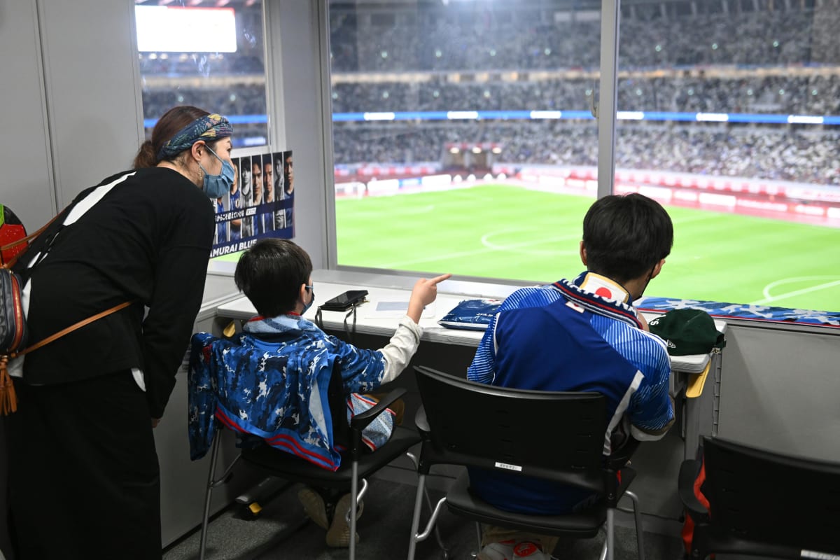 次世代を担う青いユニフォームの子どもたちが、コーチからサッカー戦術を学ぶ様子。緑のピッチでボードを囲み熱心に耳を傾ける。