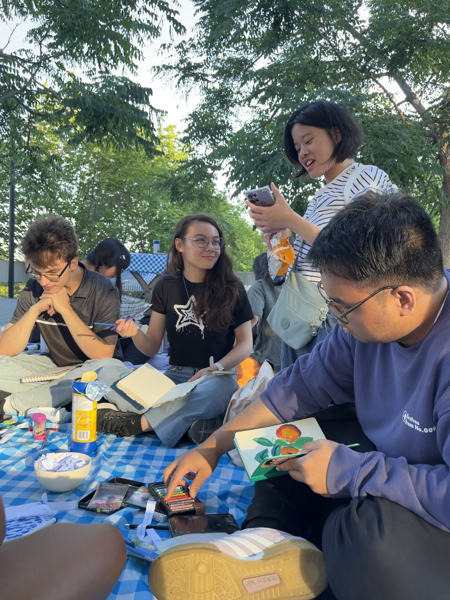 Participants at the Art Picnic sitting on the ground, near large trees, talking to each other while drawing and painting.