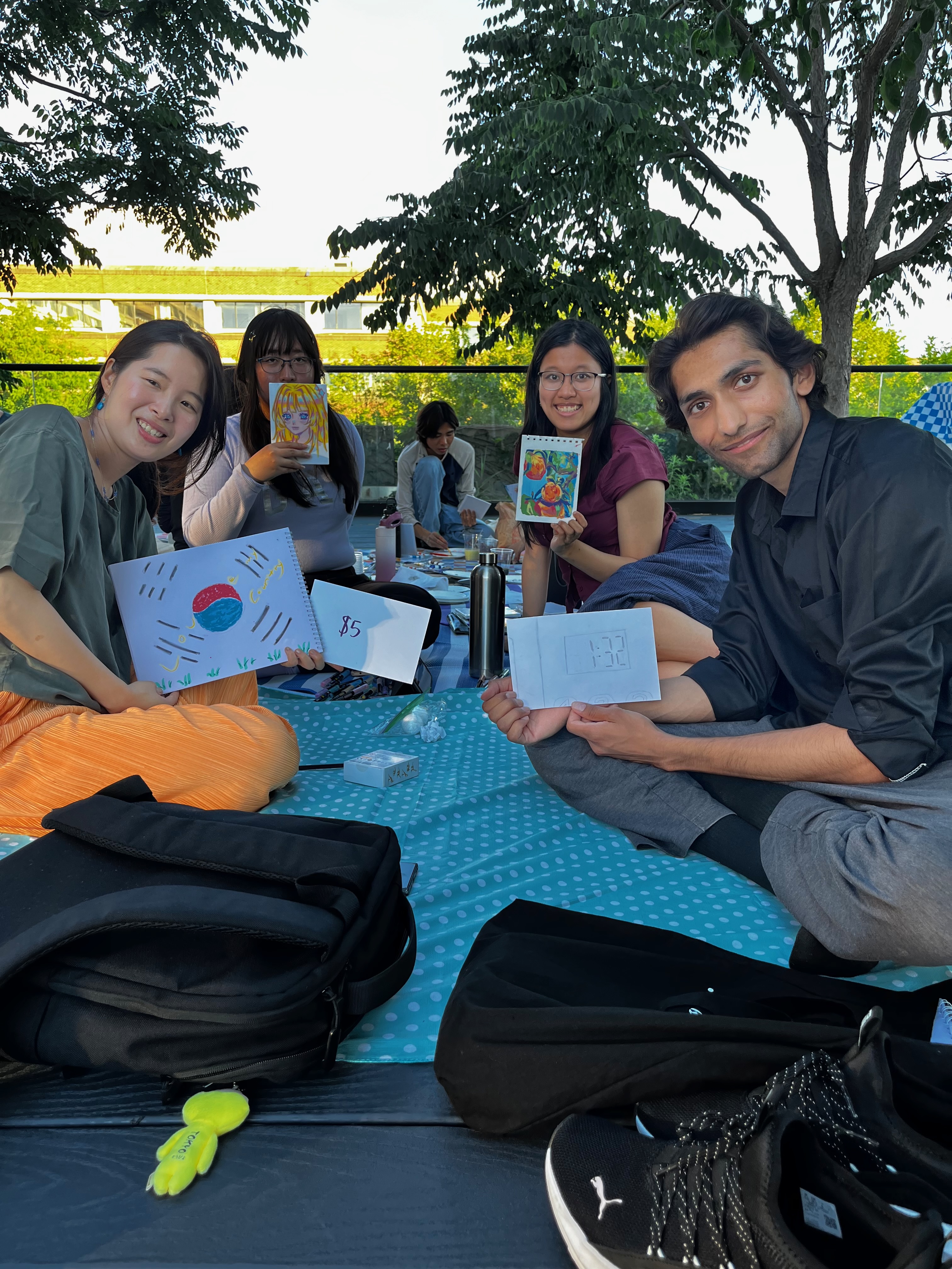 Participants at the Art Picnic sitting on the ground, near large trees, holding their artworks to the camera and smiling.