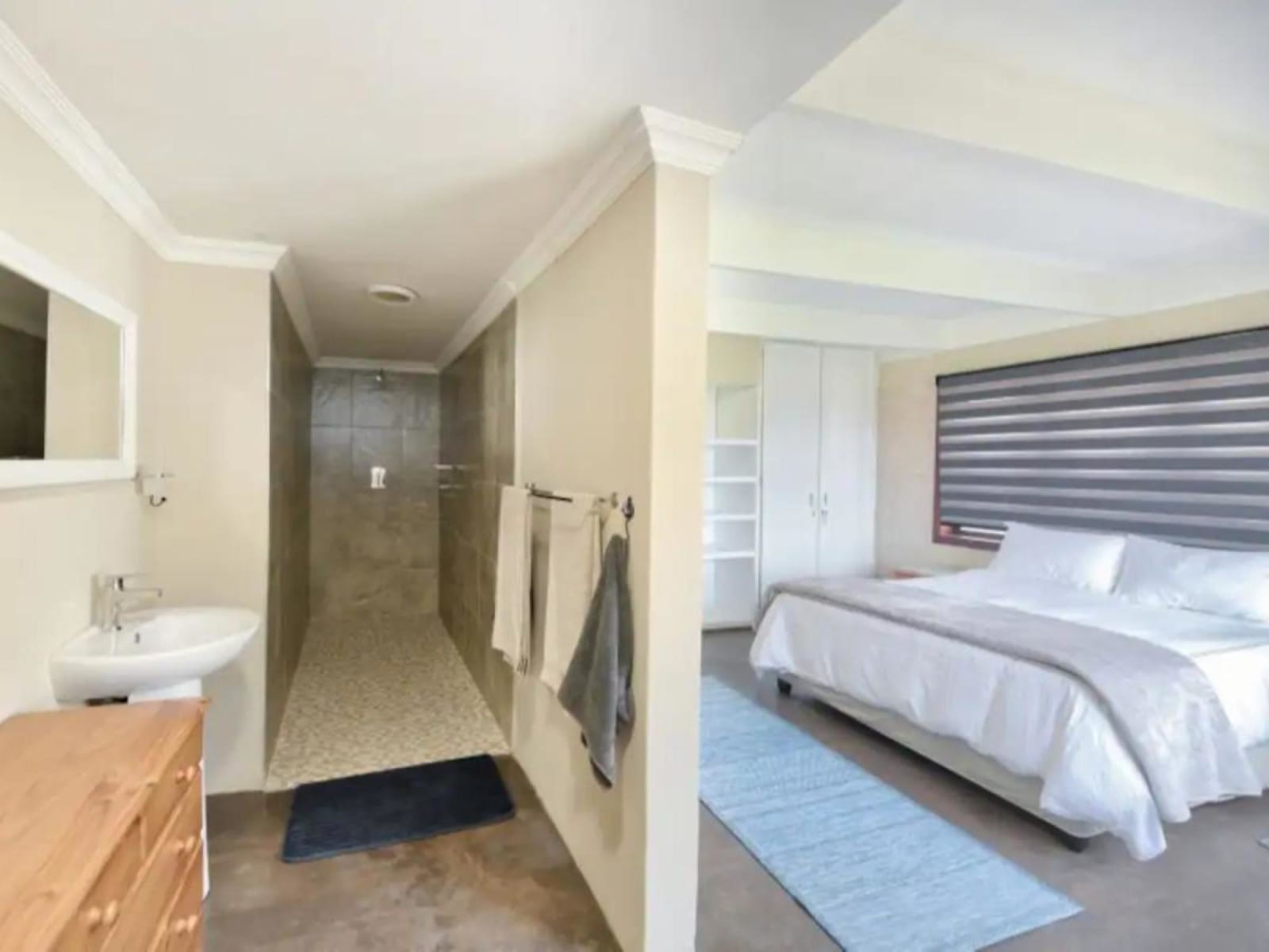 A double bedroom at Gans-te-Ver with its en-suite walk-in shower alongside — crisp white linen, blinds and a pine chest of drawers.