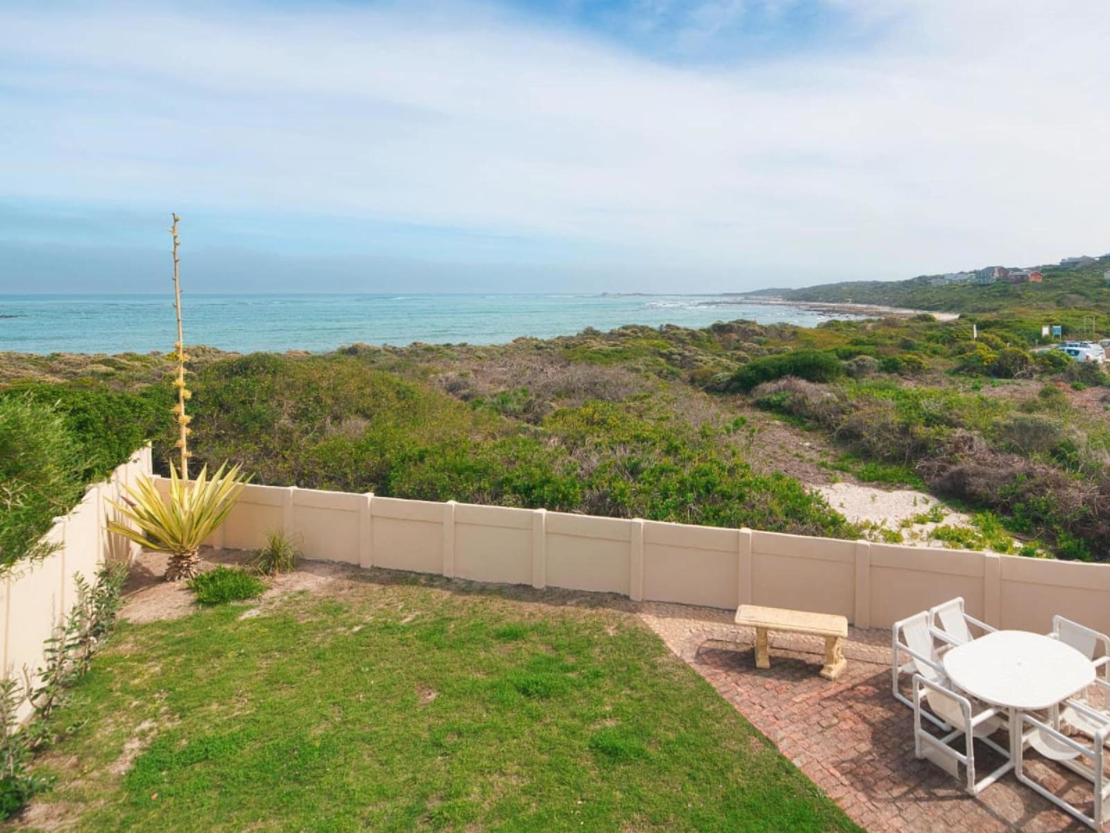The view from Gans-te-Ver — the lawn and boundary wall giving way to nature-reserve fynbos, with the beach curving away toward Struisbaai.
