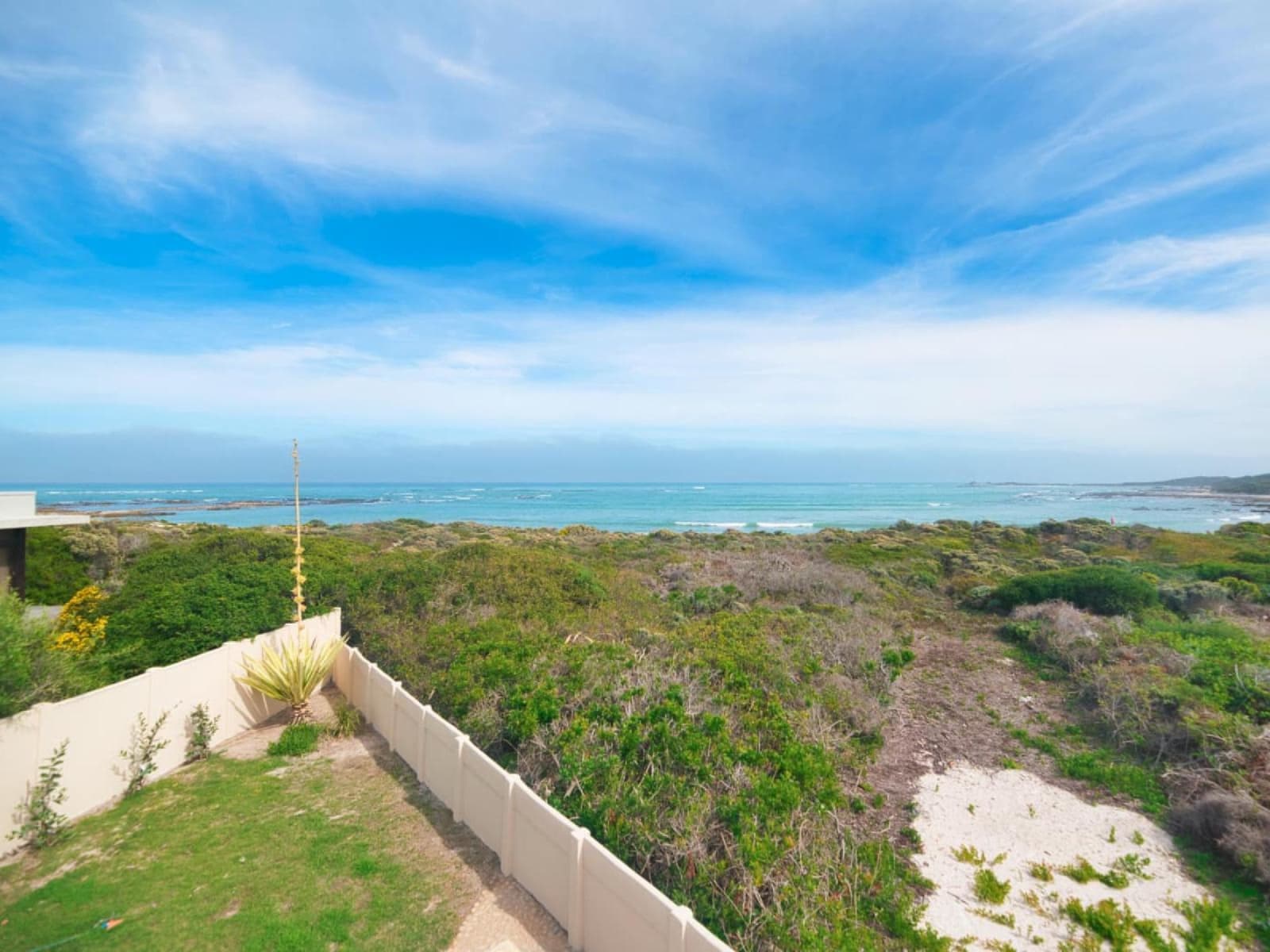 Gans-te-Ver's garden meeting the coastal nature reserve — green fynbos, white sand patches and the open sea beyond, under a big blue sky.