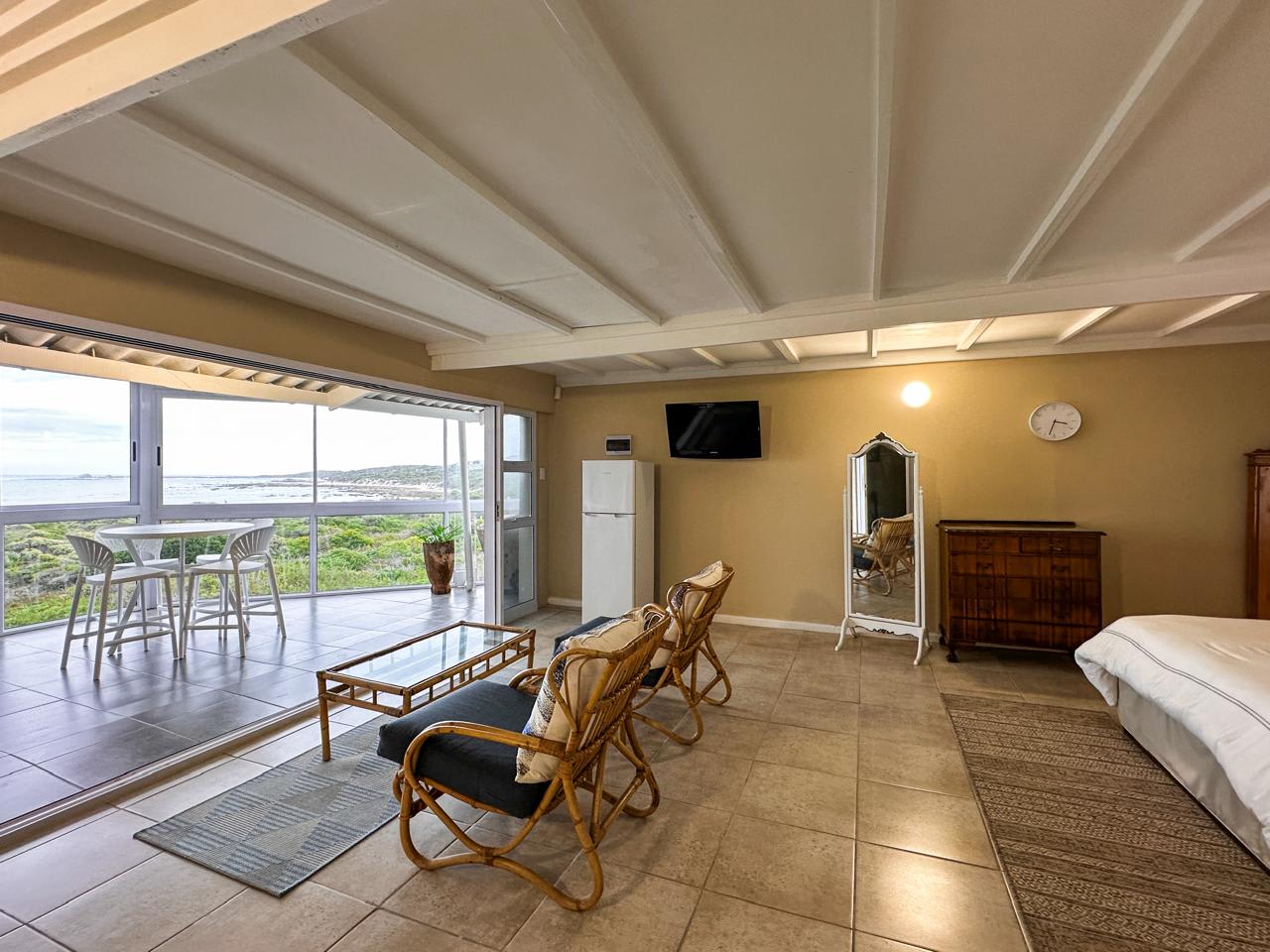 The main suite at Gans-te-Ver — bed, rattan sitting corner, TV and mirror opening onto the new glassed-in sunroom, where a bistro table sits behind sliding windows above the sea.
