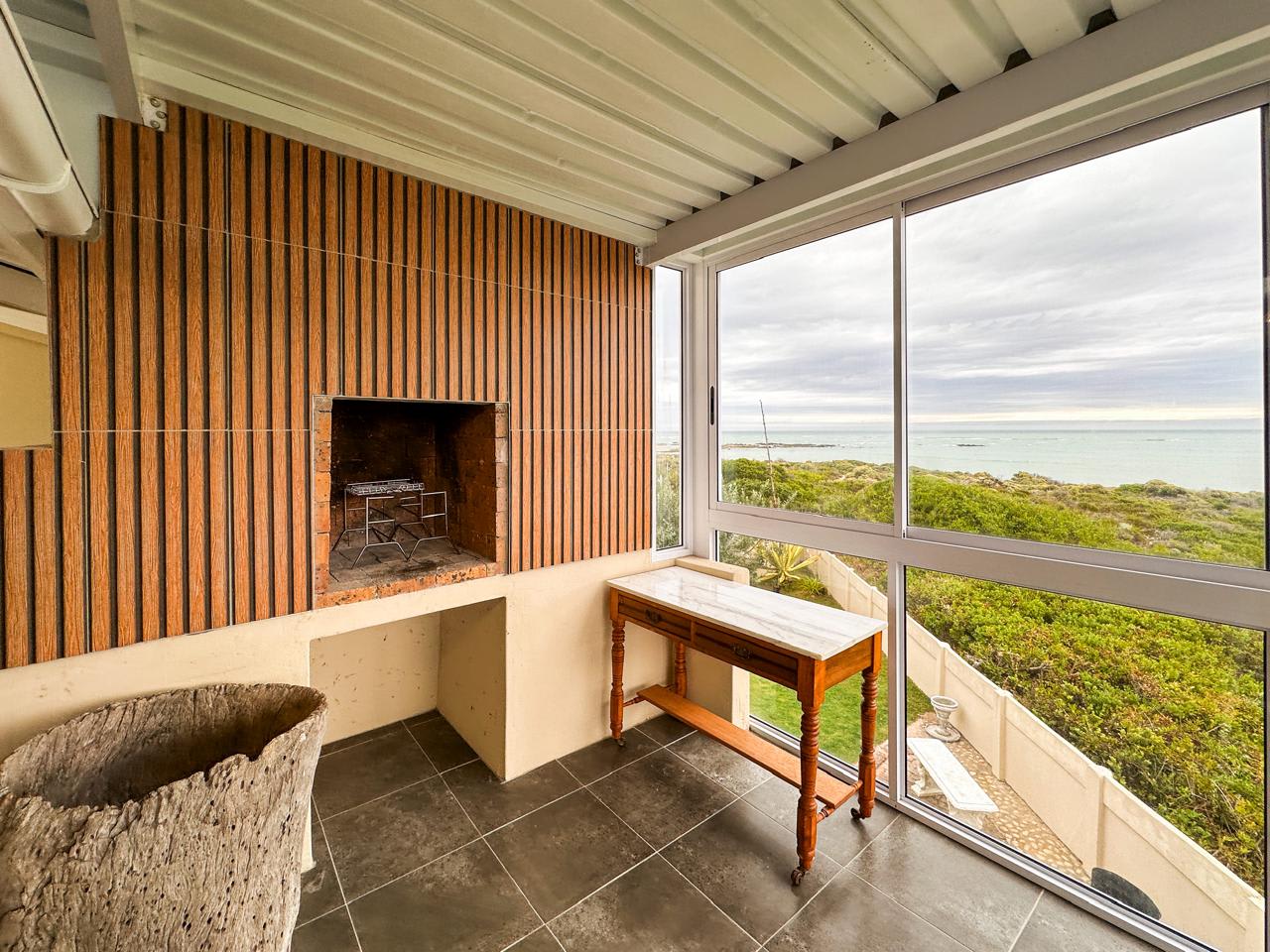 The glassed-in braai corner on Gans-te-Ver's top floor — a built-in braai against a warm timber-slat wall and a marble-topped console, with sliding glass windows opening the whole wall to the sea.