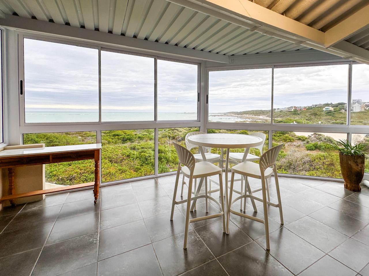 The top-floor sunroom at Gans-te-Ver — the sea-facing balcony now enclosed with frameless sliding glass windows, a bistro table and bar stools set to the view over the fynbos reserve to the Suiderstrand sea.