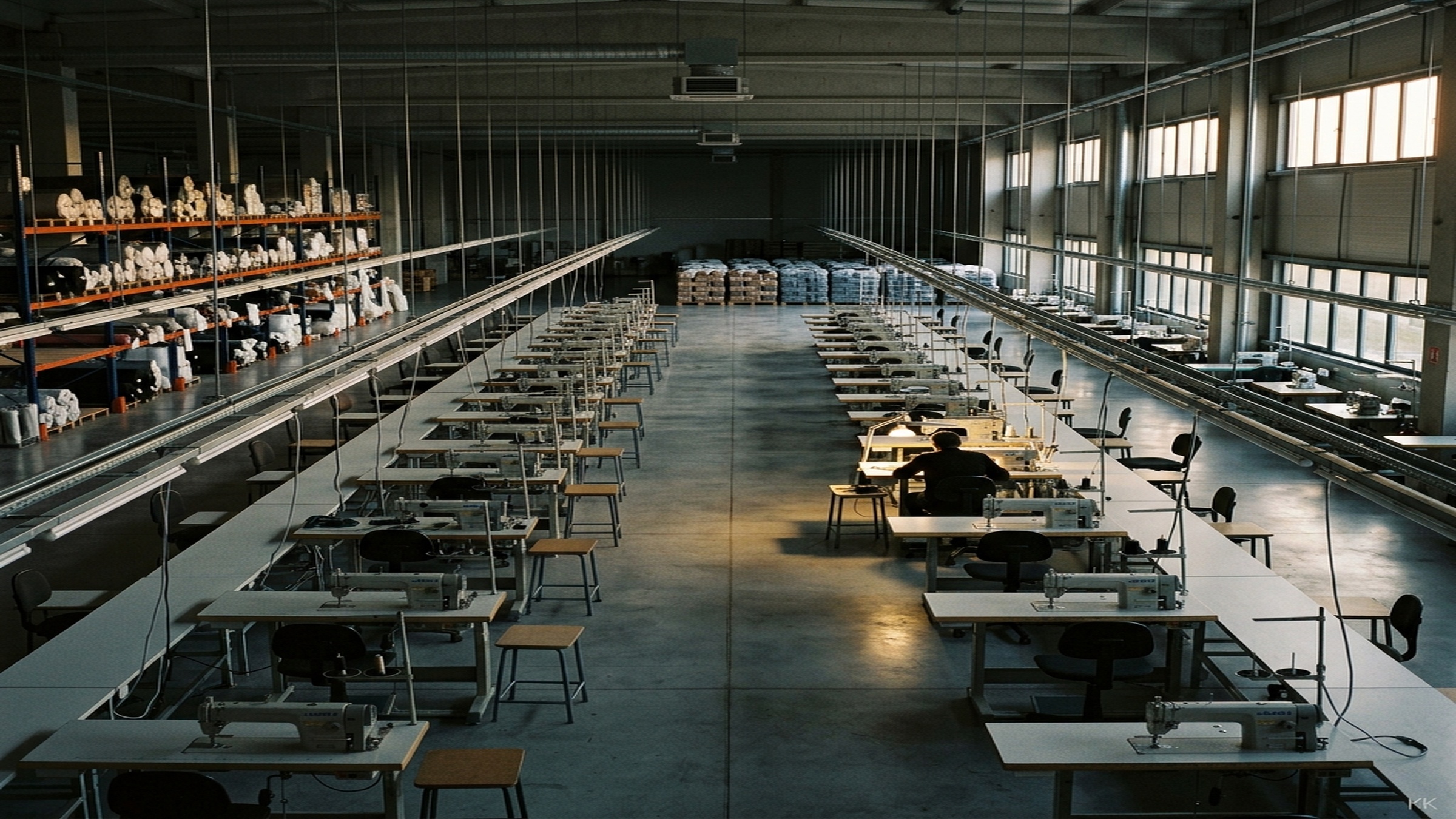 A wide documentary shot of a partially staffed apparel production floor in late afternoon, sewing stations lit but quiet, the moment before the holiday rush begins