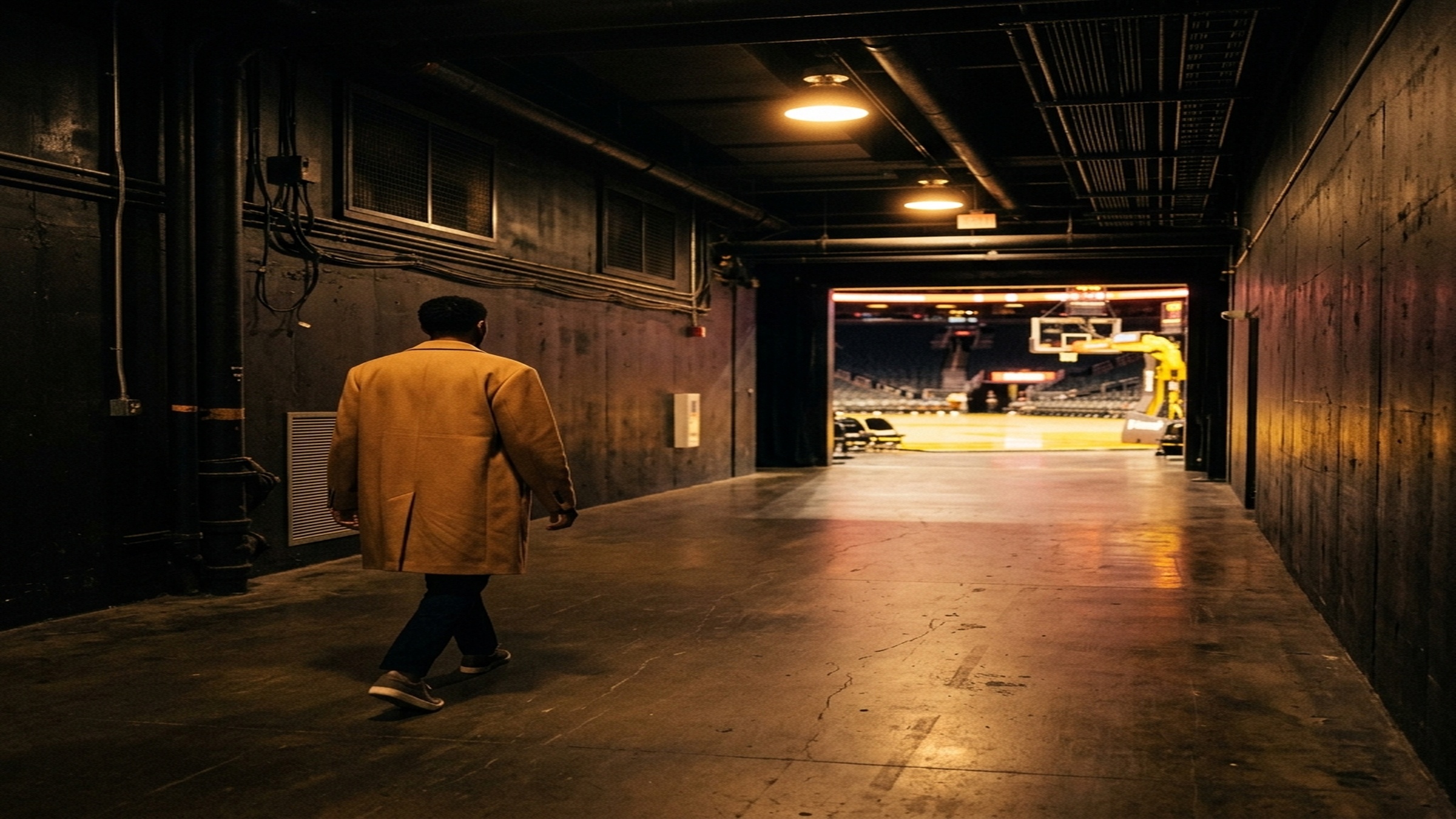 A wide, cinematic view down a darkened NBA arena tunnel — warm overhead light spilling onto a polished concrete corridor, a single figure mid-stride in a structured statement coat, photographed from behind so no face is visible, the garment's shoulder line and fabric catching the light