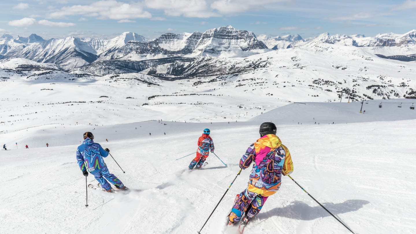 Three skiers descending an open snow slope with mountains behind them