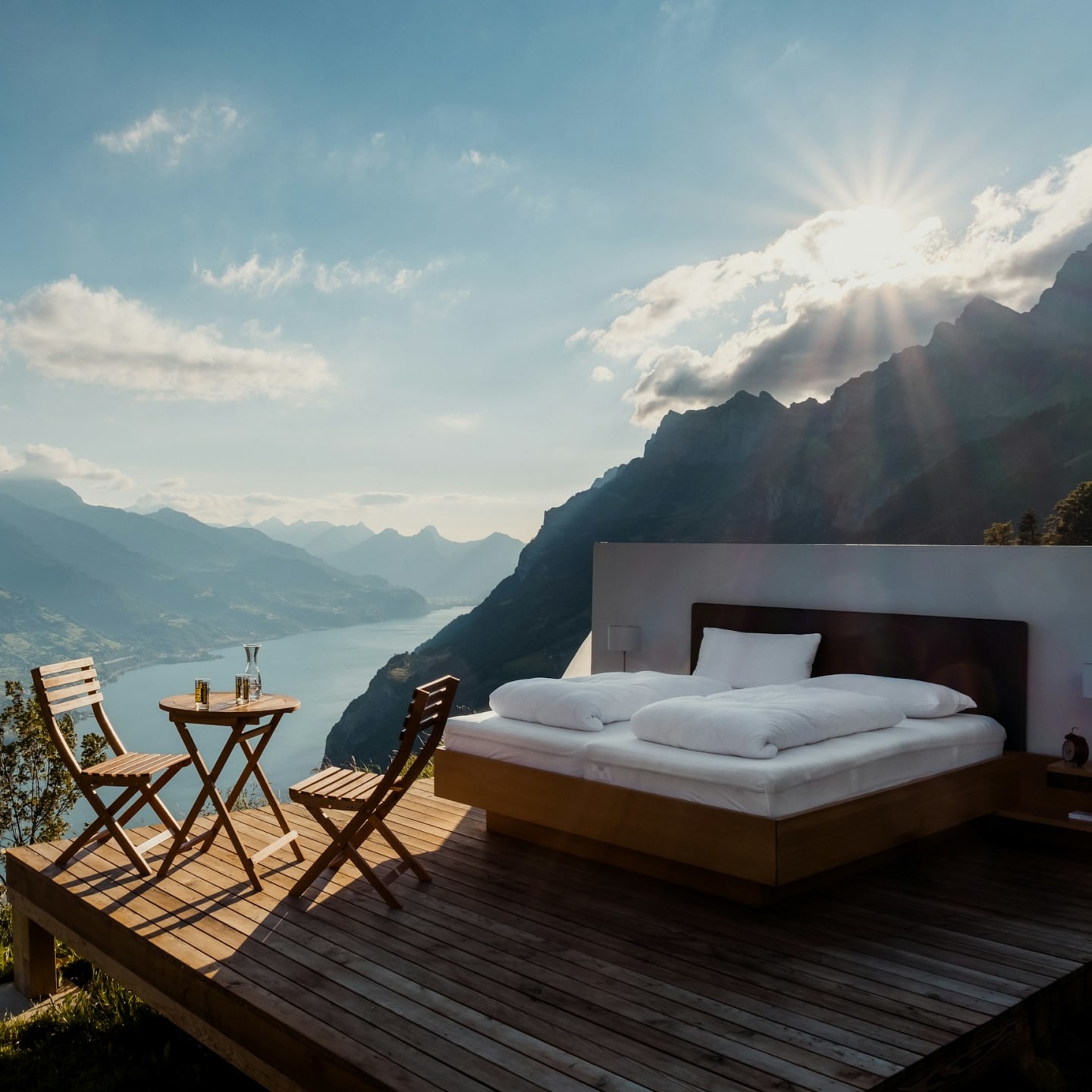 A bed and two chairs on a wooden deck overlooking a lake and mountains