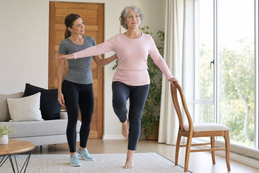 Femme de 68 ans pratiquant un exercice d'équilibre debout sur une jambe dans son salon, avec son enseignant APA à domicile