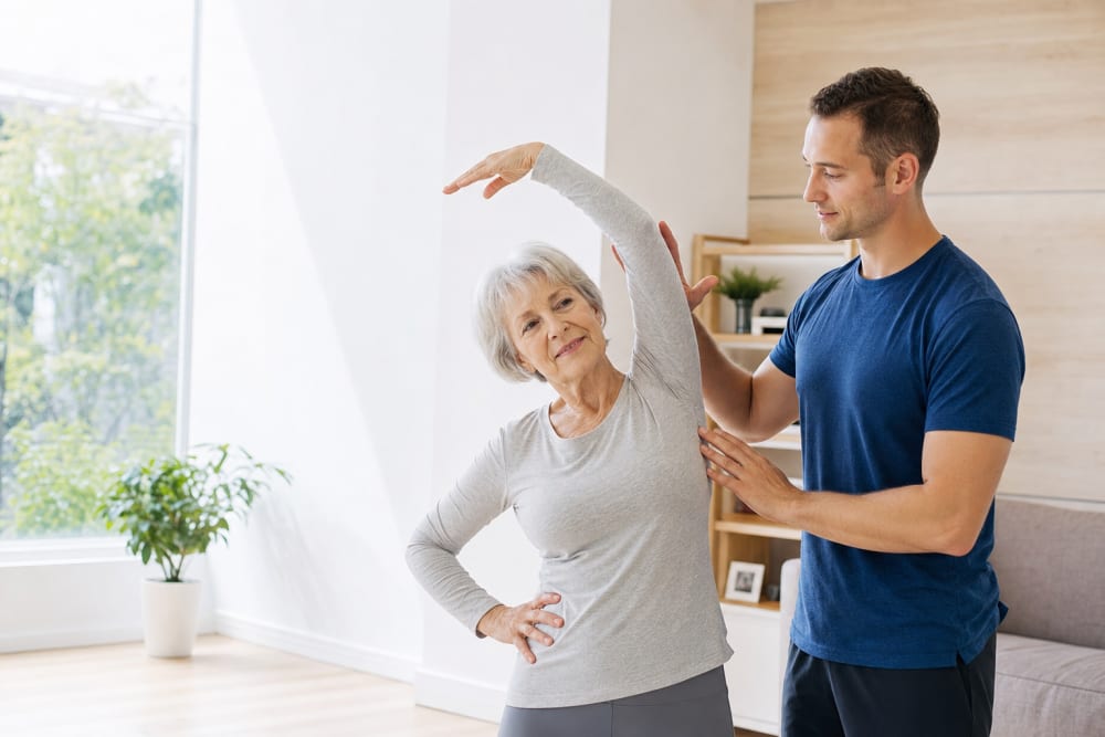Femme d'environ 72 ans réalisant un exercice d'étirement dans son salon, accompagnée d'un coach à domicile qui guide son mouvement