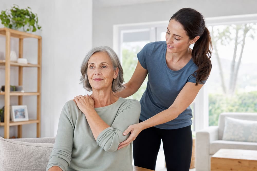 Une femme d'une cinquantaine d'années tient doucement son épaule à son domicile, accompagnée d'un coach qui guide sa posture