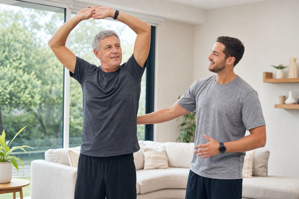 Un homme de 58 ans effectue des exercices de mobilité debout dans son salon clair, guidé par un coach sportif à domicile