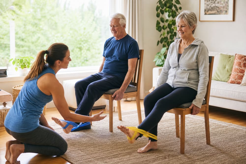 Un couple de seniors réalisant des exercices de renforcement musculaire des jambes dans leur salon, accompagnés par un enseignant APA