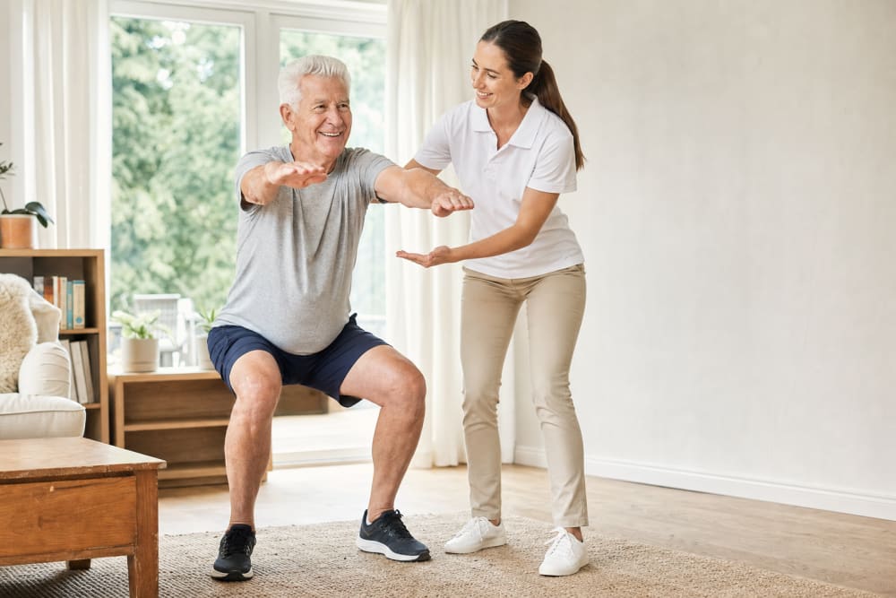 Senior homme réalisant un squat dans son salon, guidé par une coach APA qui encourage son mouvement
