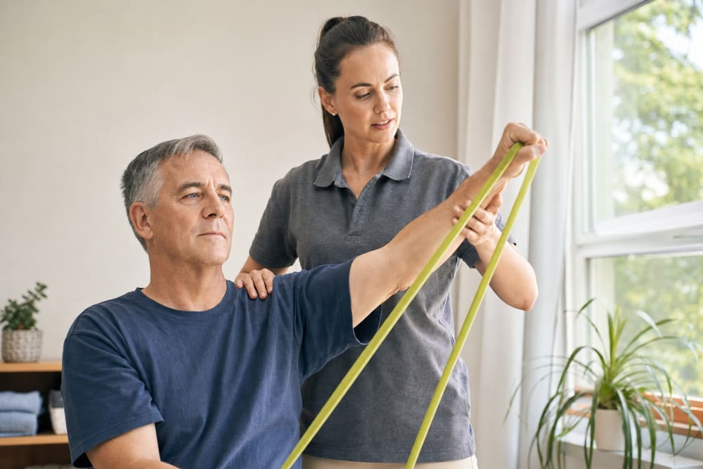 Un homme d'une cinquantaine d'années réalise un exercice de rééducation de l'épaule avec un élastique, accompagné par un enseignant APA à domicile.