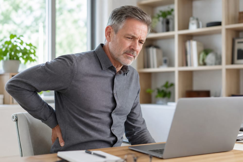 Homme d'une cinquantaine d'années en télétravail, main dans le bas du dos, illustrant les tensions dorsales causées par le stress professionnel