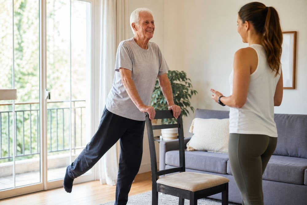 Homme âgé effectuant un exercice d'équilibre debout en s'appuyant sur une chaise, suivi par un coach APA à domicile