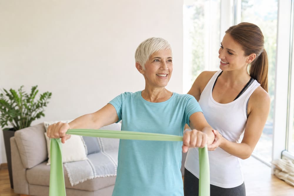 Femme de 62 ans faisant des exercices de renforcement musculaire avec un élastique dans son salon, accompagnée par un enseignant APA