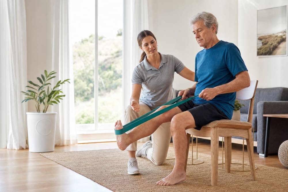 Un homme d'une soixantaine d'années réalise un exercice de renforcement avec élastique, guidé par une enseignante APA dans son salon lumineux