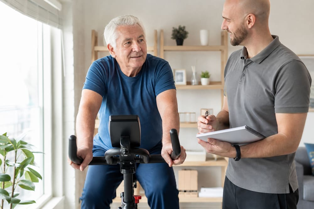 Un homme de la soixantaine pédale sur un vélo d'appartement chez lui, accompagné par son enseignant APA