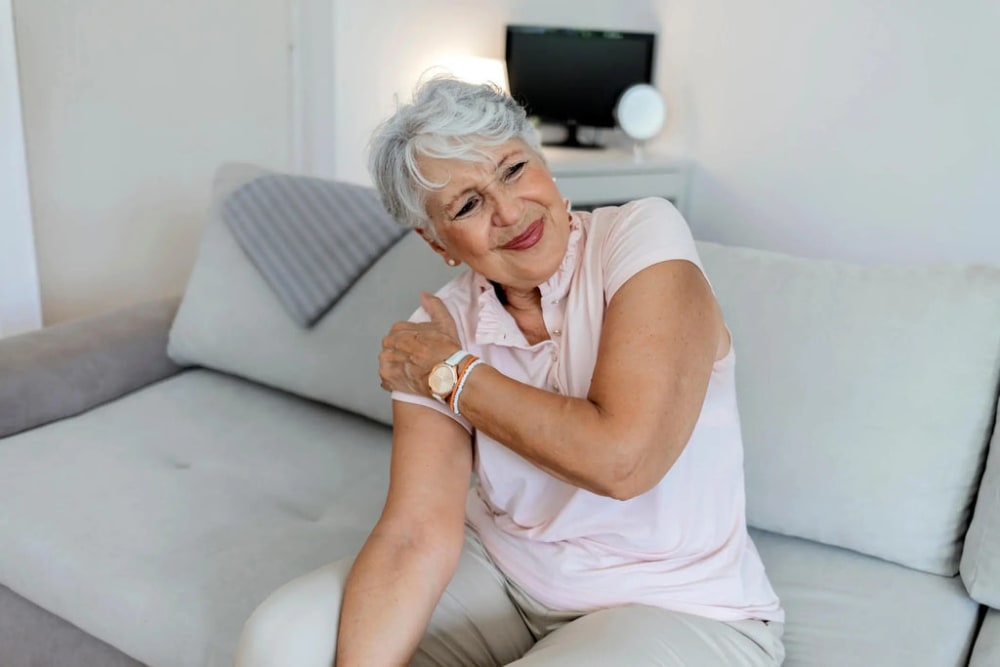 Une dame, assise sur un canapé, tient son épaule droite car elle doit avoir mal.