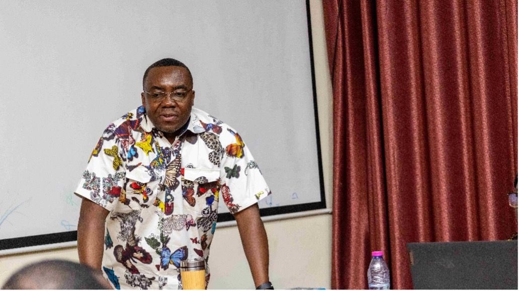 A man in a butterfly-patterned shirt stands, leaning forward, possibly presenting.