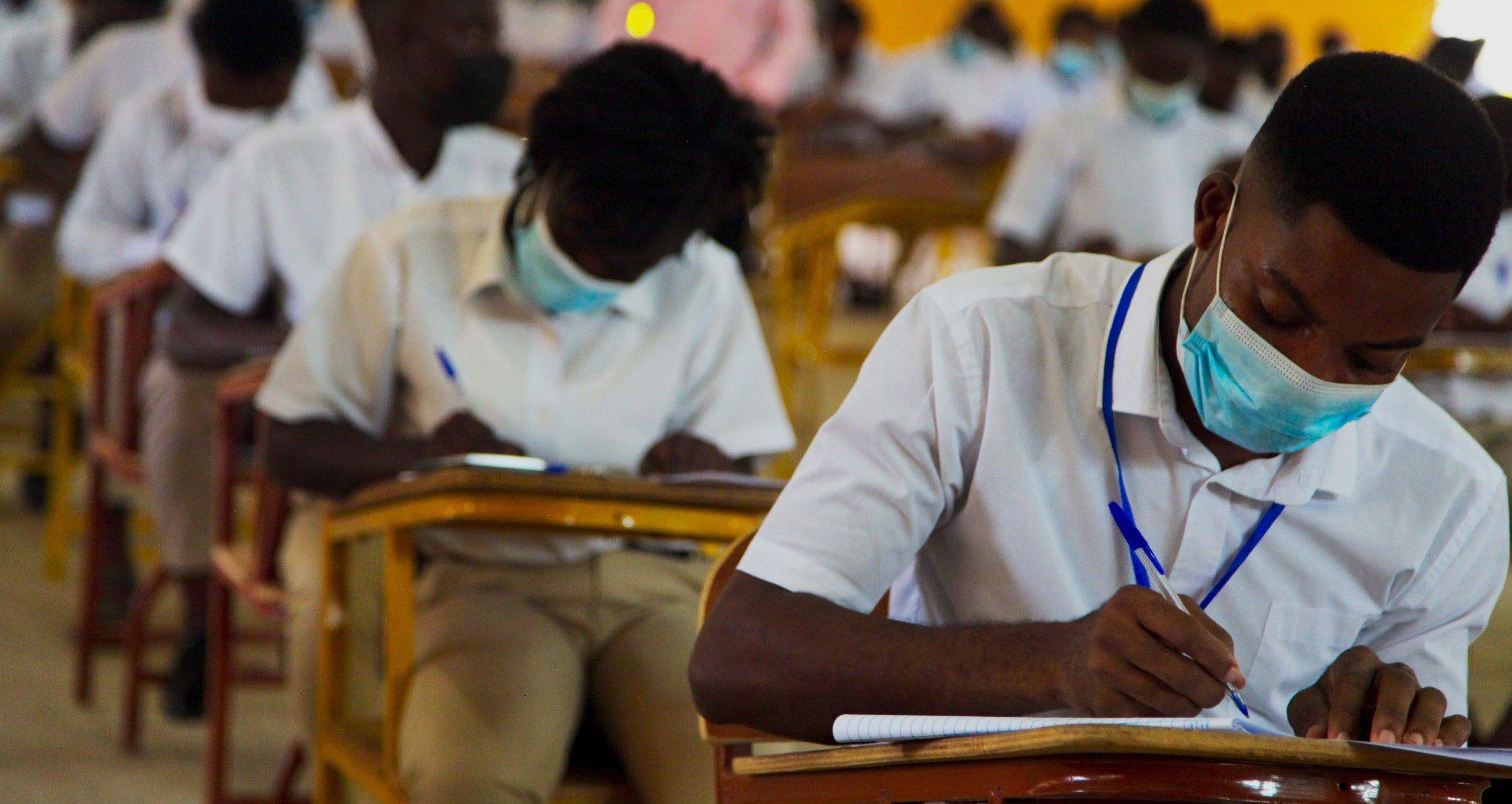 Students in masks write intently at desks during an examination in a classroom.