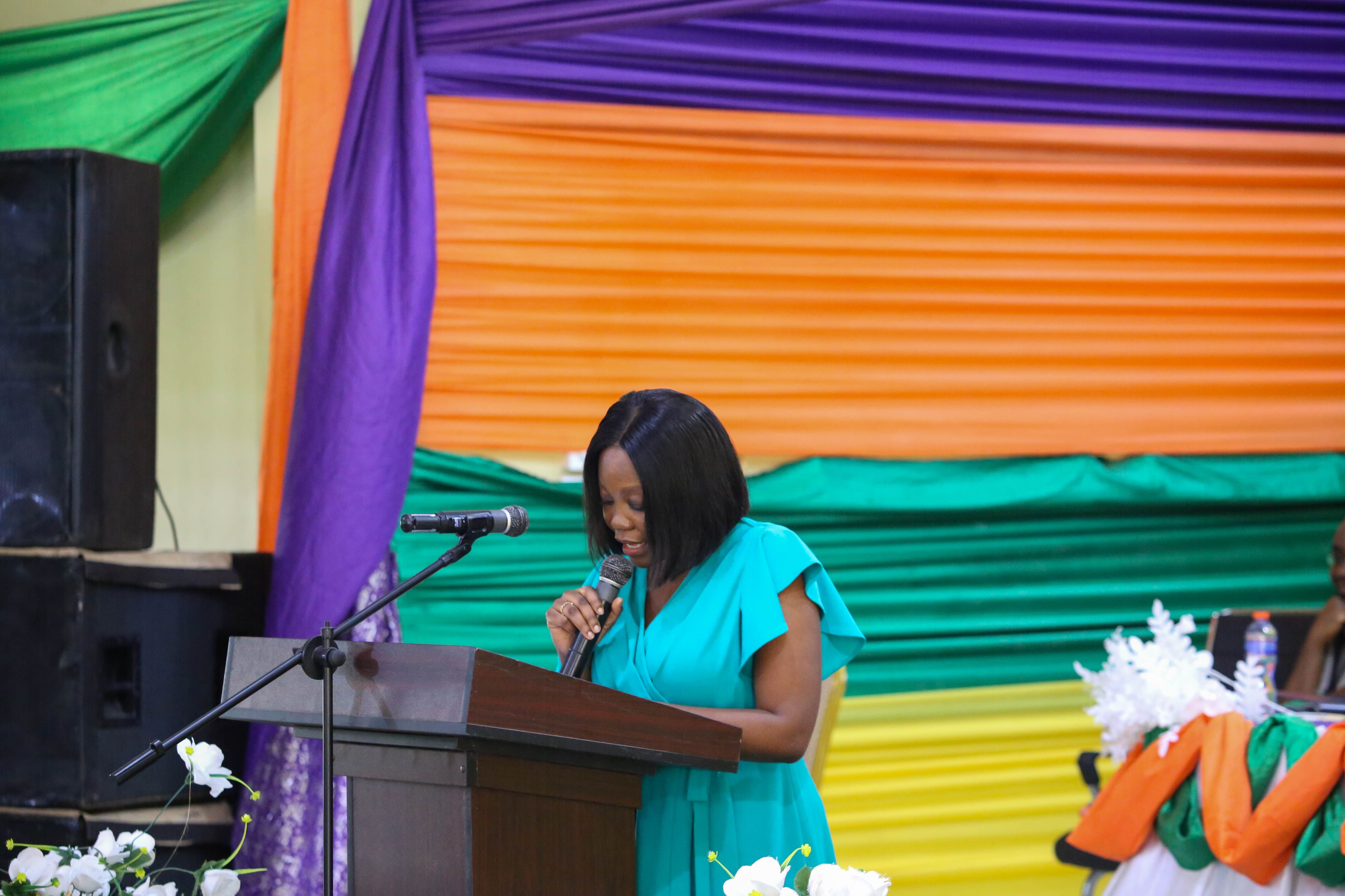 Woman in blue dress speaks at a podium with a microphone against a colorful fabric background.