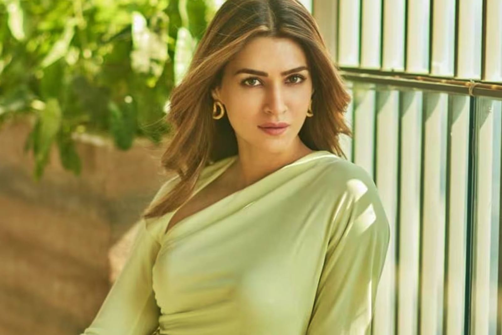 Kriti Sanon, a Bollywood actress, posing in a light lime green asymmetrical top with gold hoop earrings, looking directly at the camera with a serene expression. Her brown hair is wavy, and the background is blurred green foliage and modern architecture.