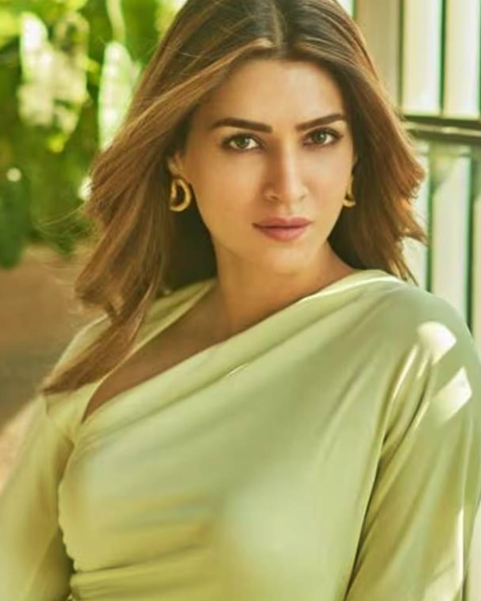 Close-up portrait of Kriti Sanon, an Indian actress, wearing a light pastel green off-shoulder top and gold hoop earrings. Her brown hair is styled in soft waves, and she looks directly at the camera with a serene and confident expression.