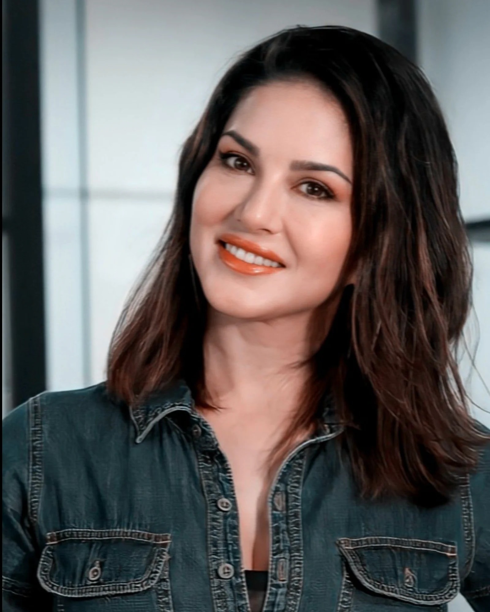 Sunny Leone smiling warmly, wearing a dark denim shirt with her dark hair styled loosely, against a blurred light background.