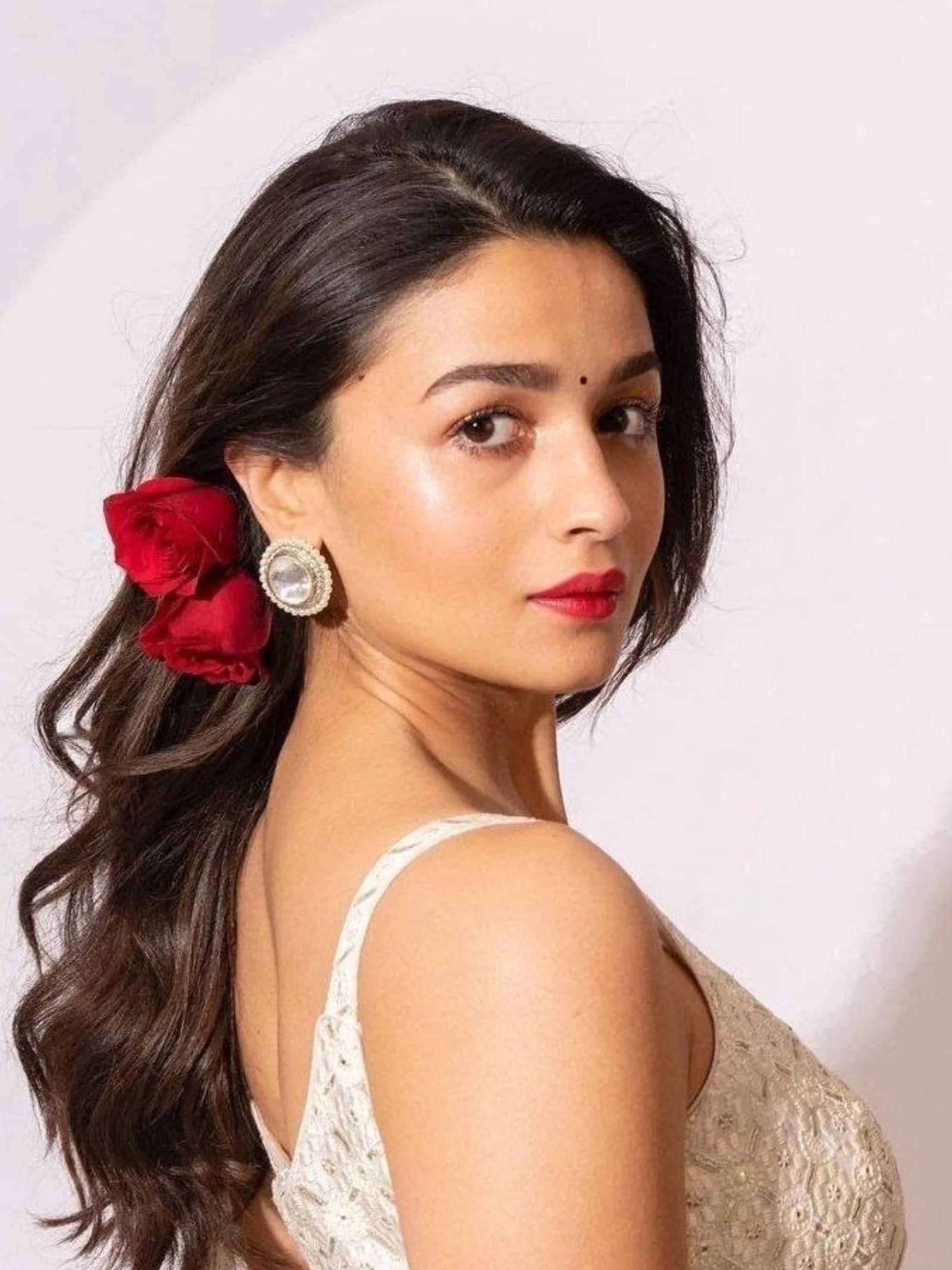 Alia Bhatt in a close-up portrait, looking over her shoulder, with dark wavy hair, two red roses, red lipstick, and a bindi, wearing a cream embroidered top.