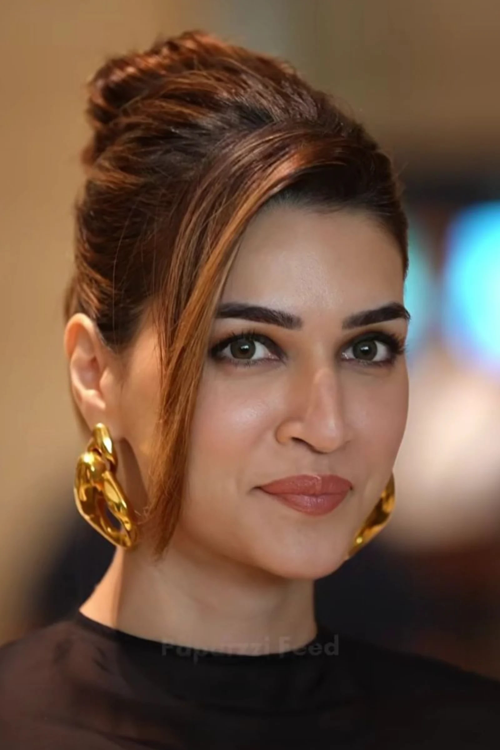 Close-up portrait of Kriti Sanon with an elegant updo, wearing large gold abstract earrings and a black top, smiling gently.