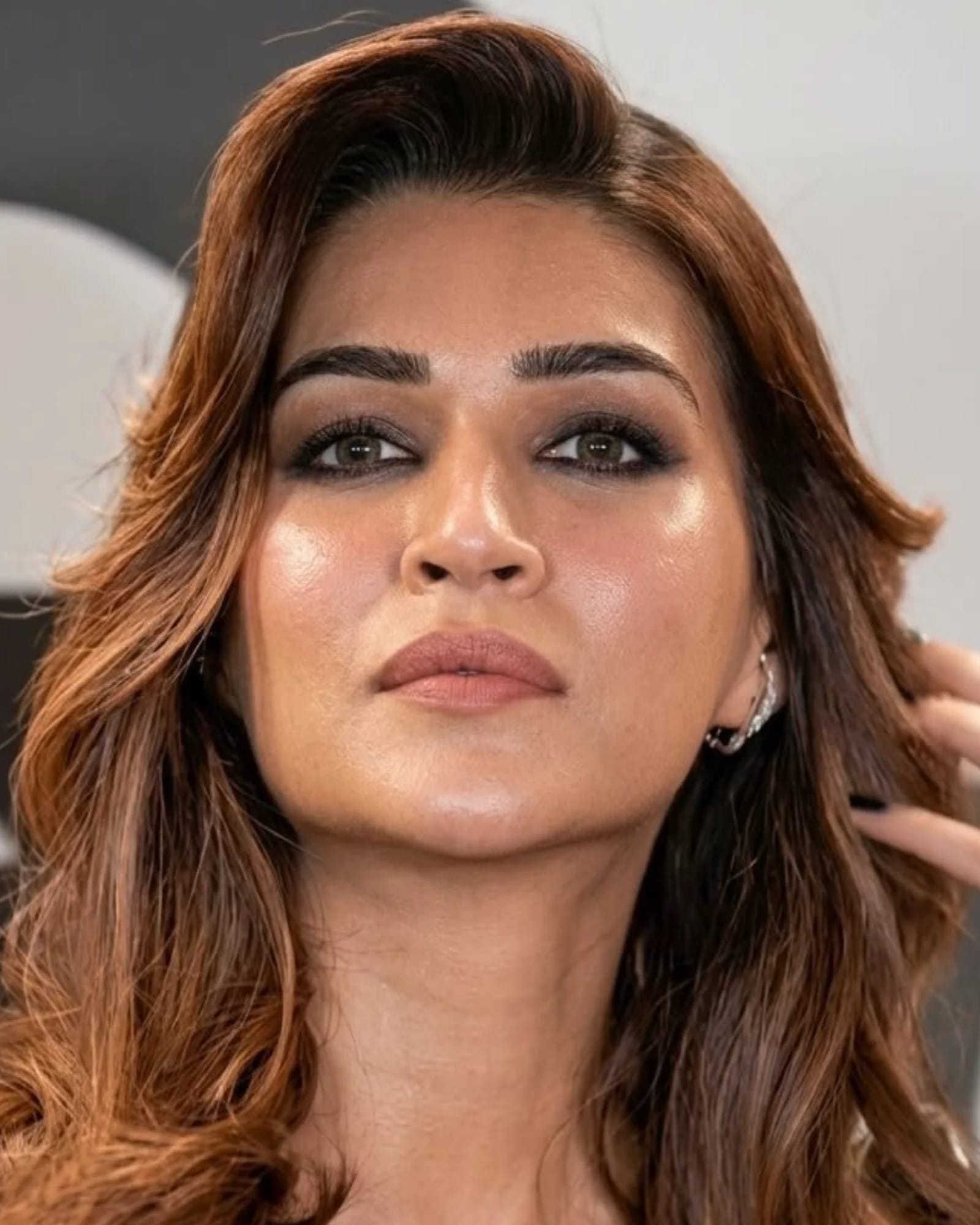 Close-up portrait of Kriti Sanon with smoky eyes, wavy brown hair, and a serene expression, gently touching her hair.