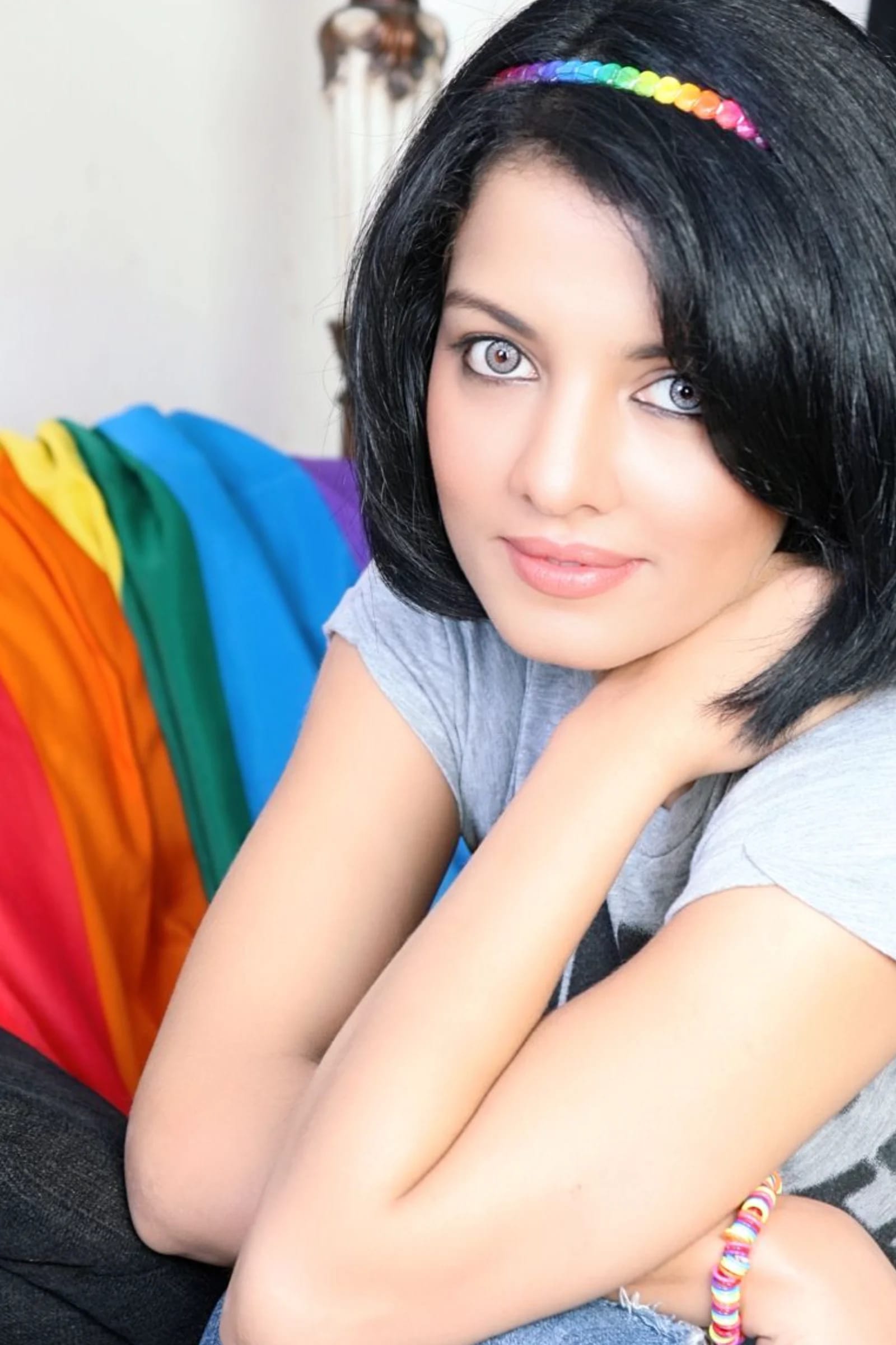 Celina Jaitly, an Indian actress, smiling gently while wearing a grey t-shirt, a rainbow headband, and a rainbow bracelet. Her dark hair is styled in a bob, and she has light-colored eyes. A rainbow-striped fabric is visible in the background.