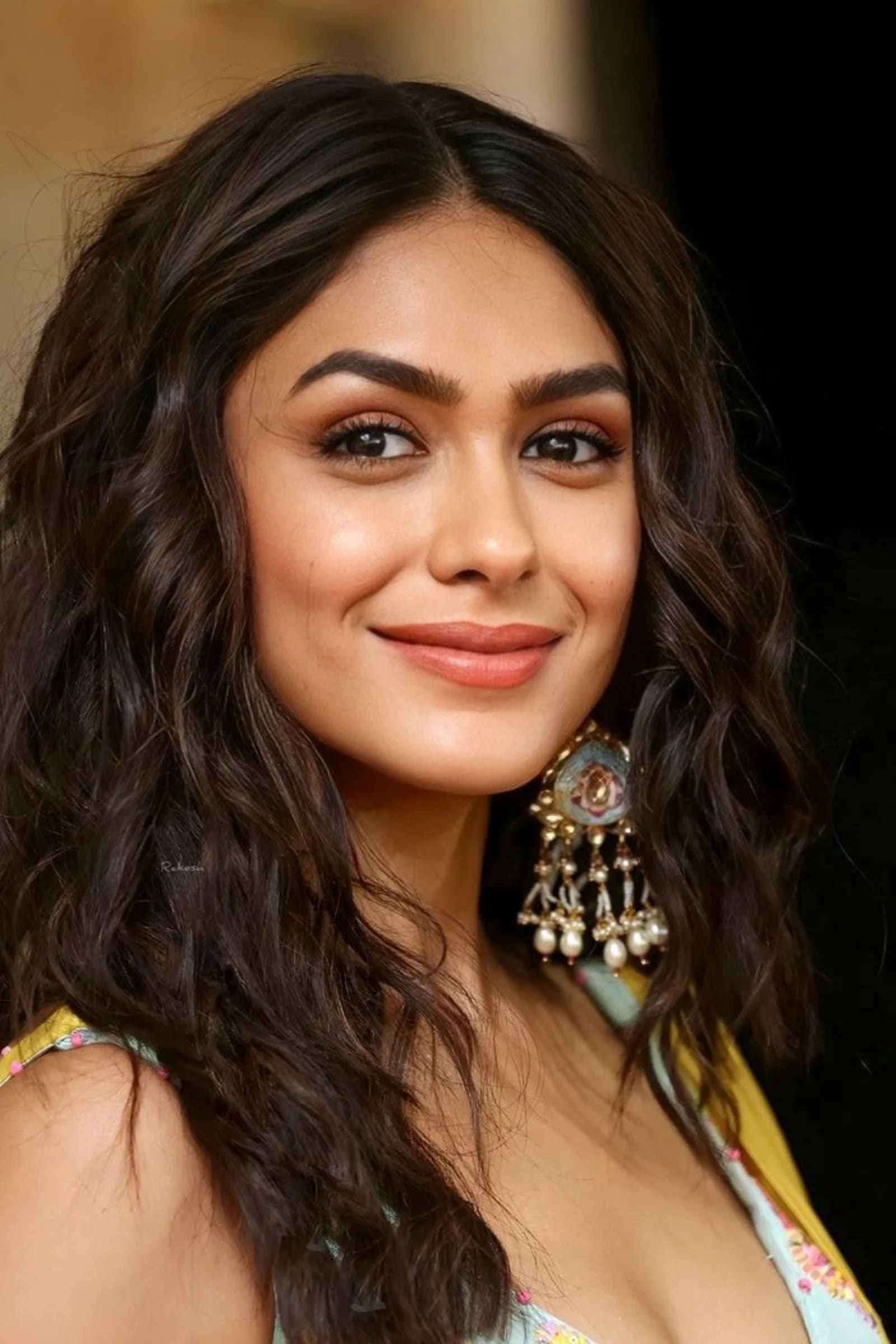 Close-up portrait of Mrunal Thakur smiling, with wavy dark hair and ornate traditional earrings.