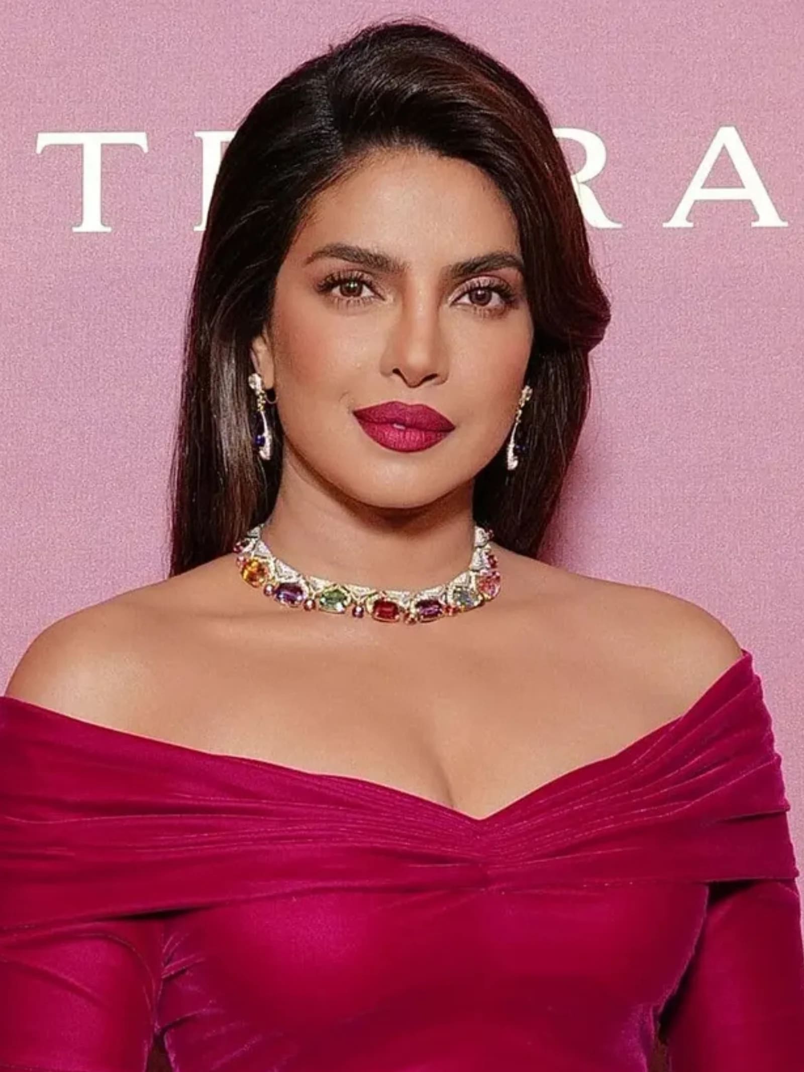 Priyanka Chopra in a magenta off-shoulder velvet dress, wearing a colorful gemstone choker necklace and drop earrings, looking directly at the camera with a confident expression.
