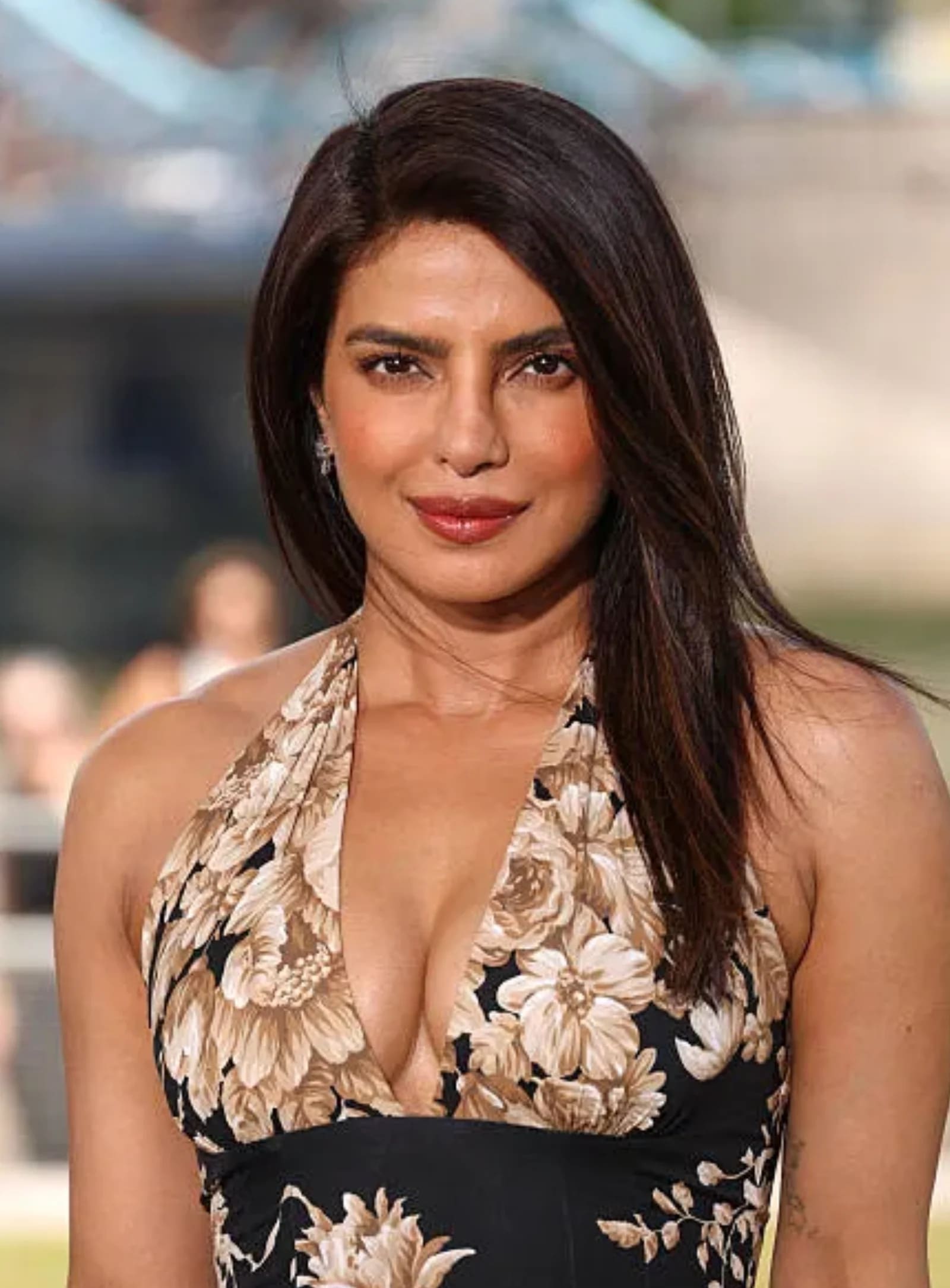 Priyanka Chopra smiling, wearing a black halter-neck dress with large beige floral patterns, with her dark hair flowing over her shoulders.