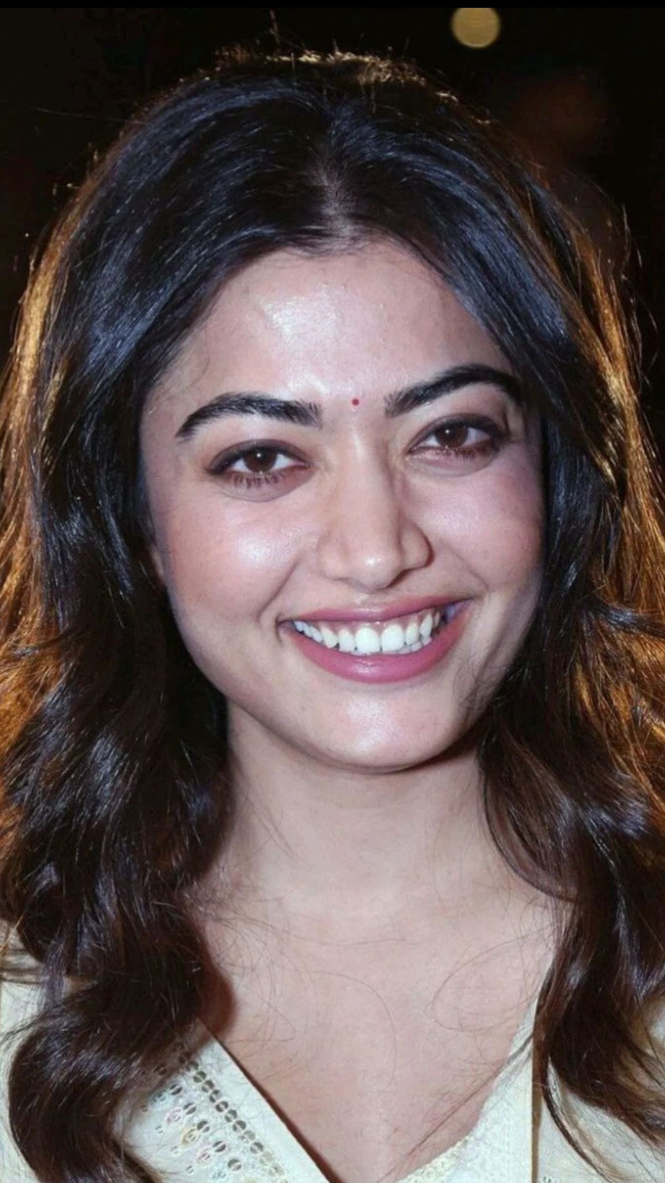 Close-up portrait of Rashmika Mandanna smiling broadly, with dark wavy hair and a small red bindi on her forehead.