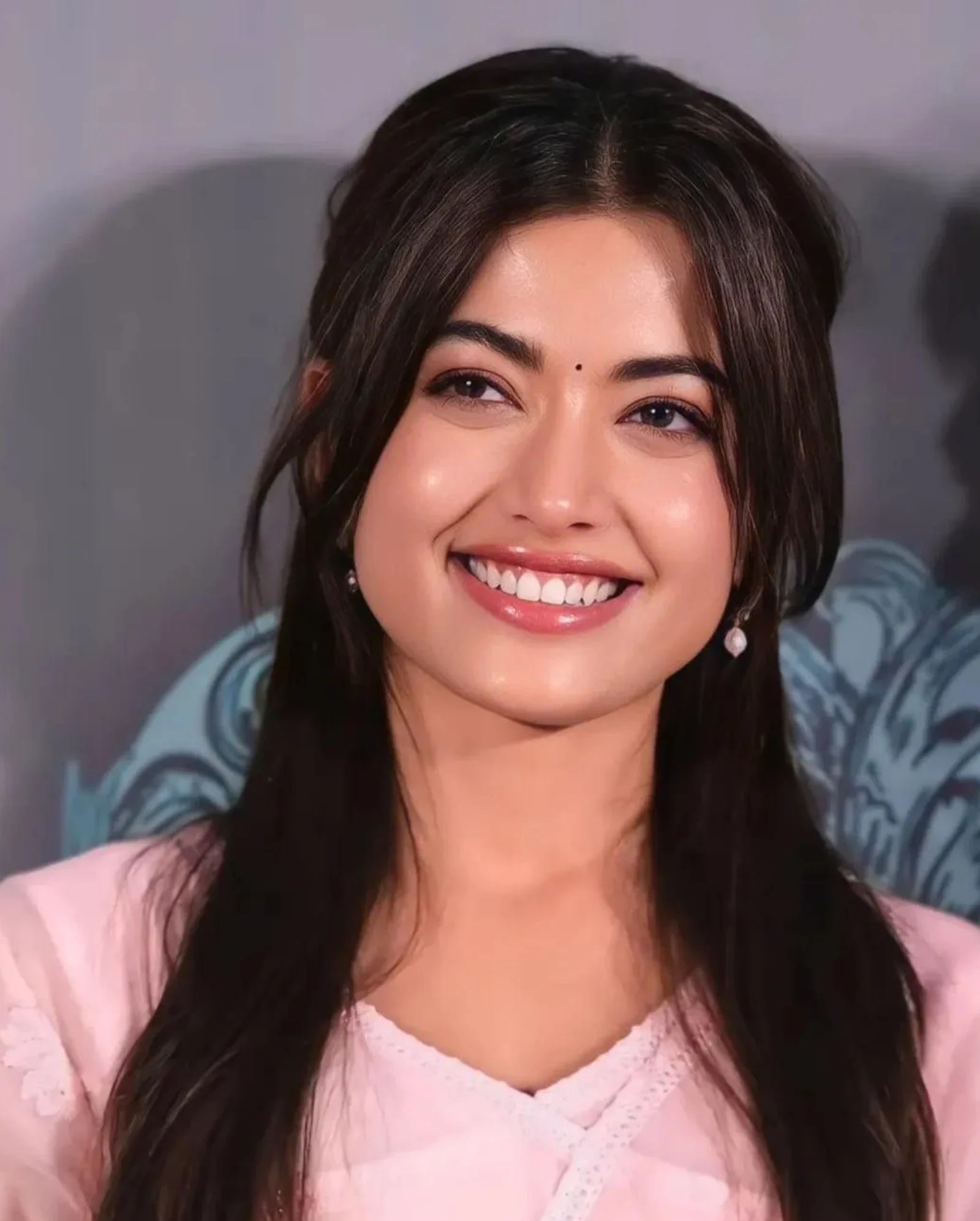 Close-up portrait of Rashmika Mandanna smiling radiantly, wearing a light pink ethnic outfit with white lace, pearl earrings, and a bindi.