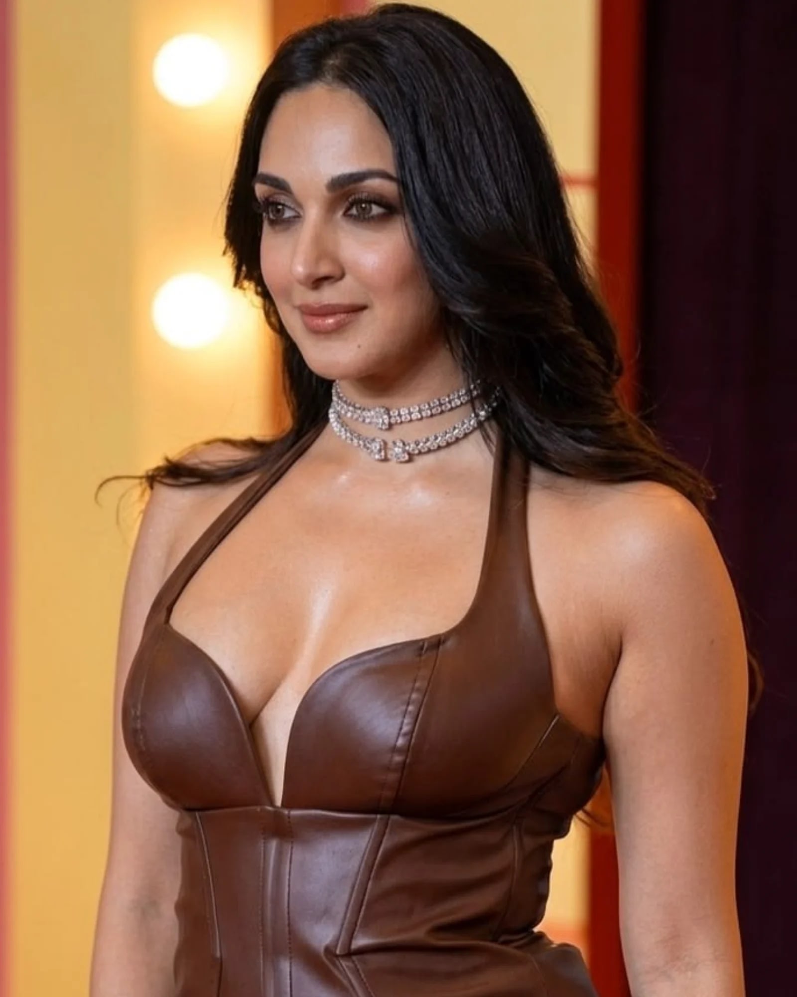 Kiara Advani, an Indian actress, wearing a brown leather halter top with a deep V-neck and two diamond choker necklaces. She has long dark wavy hair and is looking slightly to the left with a subtle, alluring smile. The background is blurred with warm yellow and dark red lighting.