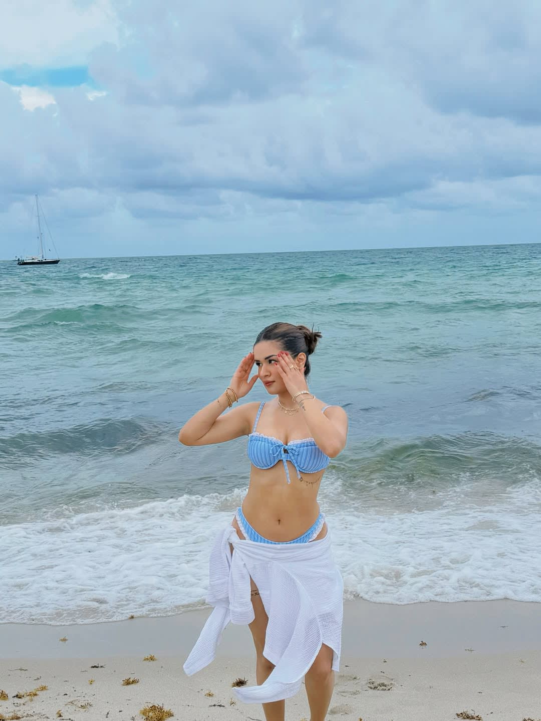 Avneet Kaur standing on a sandy beach, wearing a blue and white striped bikini and a white sarong, with her hands near her face, looking towards the ocean. A sailboat is in the background.